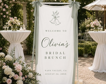 May include: A vertical white fabric banner featuring a line drawing of two bells, the text "WELCOME TO Olivia's BRIDAL BRUNCH NAPA VALLEY, CA AUGUST 24, 20XX," and sage green ribbons tied to the top corners of the wooden frame holding the banner.