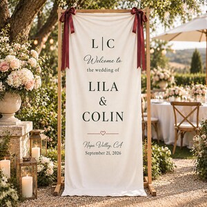 Op de afbeelding: Een verticaal, crèmekleurig stoffen welkomstbord voor een bruiloft, opgehangen aan een houten frame met donkerrode linten. De tekst luidt: L | C, Welcome to the wedding of LILA & COLIN, een klein hartsymbool, Napa Valley, CA, 21 september 2026.