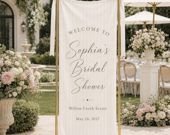 May include: A vertical cream-colored fabric banner with subtle vertical stripes, suspended from a gold metal stand with decorative bows. The text reads: WELCOME TO Sophia's Bridal Shower, Willow Creek Estate, May 24, 2027, with a small heart icon.