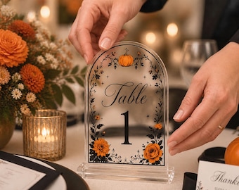 May include: An arched, frosted acrylic table number sign featuring the text "Table 1" in black, decorated with a printed illustration of a pumpkin at the top and orange flowers with black foliage along the sides, set on a clear acrylic base.