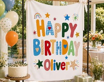 May include: A white fabric birthday backdrop featuring the text Happy Birthday Oliver in colorful, patterned letters. The design includes various shapes like stars, hearts, and a rainbow, suspended on a black metal frame with white ribbons.