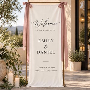 May include: A vertical cream-colored fabric wedding welcome sign hanging from a gold metal frame. The sign features black text reading Welcome to the wedding of Emily & Daniel, September 20, 2025, Napa Valley, California. Dusty rose ribbons are tied at the top.