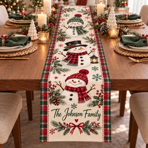 May include: A cream-colored table runner featuring three illustrated snowmen, pine branches, pinecones, and red berries. The runner has a red and green plaid border and includes the text The Johnson Family above a red bow and heart.