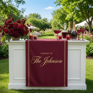 May include: A white bar table draped with a deep red runner featuring gold text that reads "CHEERS TO The Johnson". The table holds a large bouquet of dark red roses, two glass bottles, two champagne flutes, two coupes, and two red candles.