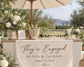 May include: A beige linen table runner featuring the black printed text "They're Engaged," the names "Olivia & Jackson," the date "August 16, 2026," and a small heart icon. The runner is draped over a white bar with champagne and glasses.