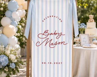 May include: A vertical rectangular sign featuring light blue and white vertical stripes, adorned with two large light blue satin bows at the top corners. The text reads CELEBRATING Baby Mason and the date 08.16.2027 in a dark red script and print.
