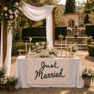 May include: A rectangular table covered with a white tablecloth featuring a cream-colored runner with the black text Just Married. The table is decorated with white floral arrangements, candles, and glassware, positioned under a wooden arch with white fabric.
