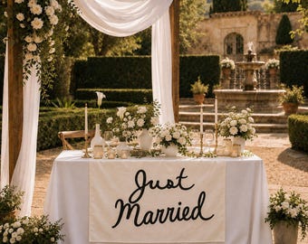 May include: A rectangular table covered with a white cloth featuring a banner that reads Just Married in black script. The table is decorated with white floral arrangements, candles, and glassware, positioned under a wooden arch with white fabric.