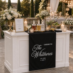 May include: A white bar counter draped with a black runner featuring the white text Cheers to The Whitmores Est. 2026. The bar holds three glass decanters on a gold tray, a framed signature drink sign, white floral arrangements, and candles.