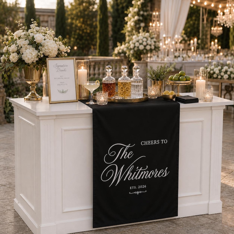 May include: A white bar counter draped with a black runner featuring the white text Cheers to The Whitmores Est. 2026. The bar holds three glass decanters on a gold tray, a framed signature drink sign, white floral arrangements, and candles.