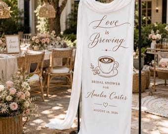 May include: A white fabric banner hanging on a black metal stand, featuring brown text that reads Love is Brewing, Bridal Shower for Amelia Carter, and August 14, 2026, with a graphic illustration of a coffee cup with latte art in the center.