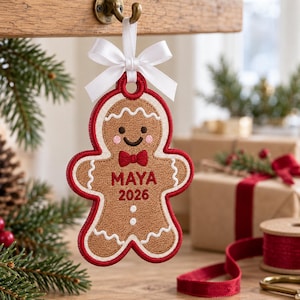 May include: A gingerbread person ornament featuring brown fabric with red stitched edges, a white satin bow, and white icing details. The centre is embroidered with the name MAYA and the year 2026 in red text, with two white buttons below.