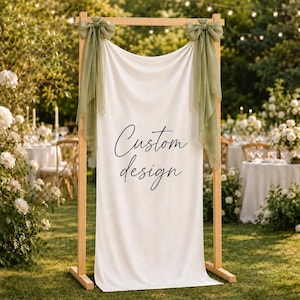 May include: A rectangular wooden frame stand supporting a white fabric backdrop. The fabric features the black text Custom design in a cursive script. Two sage green sheer fabric bows are tied to the top corners of the wooden frame structure.