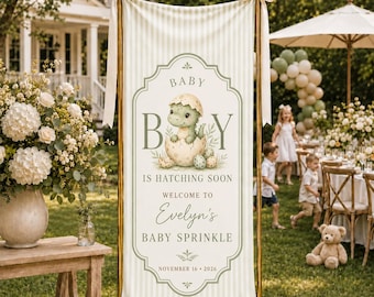 May include: A vertical banner with a light cream and white striped pattern, featuring a central decorative frame with a green dinosaur hatching from an egg. The text reads: BABY BOY IS HATCHING SOON, WELCOME TO Evelyn's BABY SPRINKLE, NOVEMBER 16, 2026.