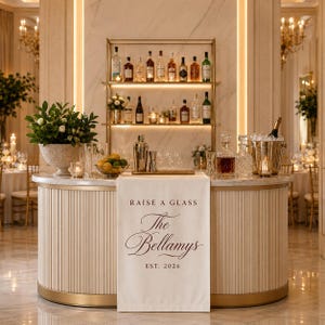 May include: A cream-colored fabric bar runner draped over a curved bar counter. The runner features dark brown text that reads: RAISE A GLASS, The Bellamys, EST. 2026. The bar is stocked with various glass bottles, a metal shaker, and a bowl of fruit.