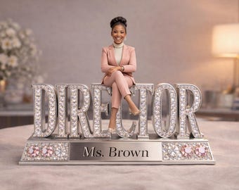 May include: A desk ornament featuring a figurine of a person in a pink suit sitting on top of large, silver, crystal-encrusted letters spelling DIRECTOR. The base is silver with crystal accents and the engraved text Ms. Brown in the center.