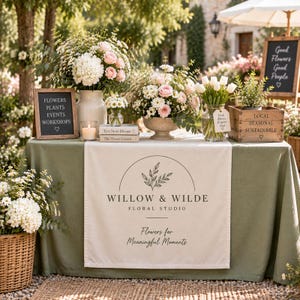 Puede incluir: Una mesa cubierta con un mantel verde salvia y un camino de mesa blanco con el texto Willow & Wilde Floral Studio Flowers for Meaningful Moments. La mesa muestra arreglos florales en jarrones, una vela, libros y letreros de madera con texto.