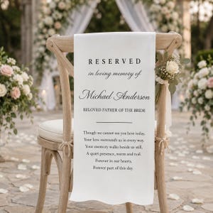May include: A white fabric sign draped over a light wood cross-back chair. The sign features black text reading: RESERVED in loving memory of Michael Anderson, BELOVED FATHER OF THE BRIDE, followed by a poem about memory and love.