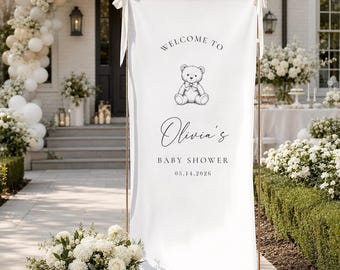 May include: A white baby shower welcome sign with the text "Welcome to Olivia's Baby Shower 03.14.2026" and a teddy bear illustration. The sign is held by a gold metal frame and decorated with white bows. The background includes white flowers and balloons.