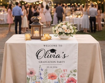 May include: A table set for a graduation party, featuring a floral runner with the text "Welcome to Olivia's Graduation Party 05.18.2026." The table is decorated with a lantern, candles, and a floral arrangement.