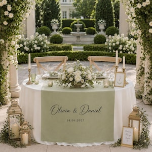 May include: A round wedding table with a sage green runner featuring the printed text Olivia & Daniel and the date 14.06.2027. The table is set with white plates, glassware, gold candle holders, and a central floral arrangement of white roses.