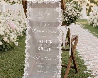 May include: A sheer white fabric memorial sign draped over a wooden folding chair, featuring white lace trim and printed text: A Seat Reserved in Heaven, FATHER OF THE BRIDE, Michael Anderson, Forever in our hearts, and October 12, 2025.