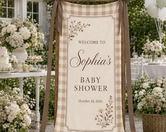May include: A vertical rectangular fabric banner with a beige and white buffalo check border, featuring the text "WELCOME TO Sophia's BABY SHOWER October 18, 2026" in dark brown, accented with botanical illustrations and tied to a wooden frame with brown ribbons.