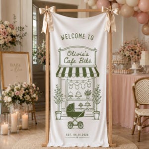 Peut inclure: Une bannière en tissu blanc suspendue à un cadre en bois avec des rubans dorés, illustrée en vert d'une devanture de café, de bodies, d'une poussette et du texte "WELCOME TO Olivia's Cafe Bébé" avec la date "EST. 14.06.2026".