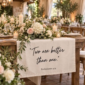 May include: A cream-colored linen table runner featuring the black printed text "Two are better than one." and "Ecclesiastes 4:9" draped over a wooden table, decorated with a floral centerpiece of white hydrangeas, pink roses, and candles.