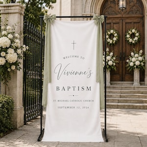 Pode incluir: Uma faixa vertical de tecido branco pendurada num suporte de metal preto, com laços de tecido verde sálvia nos cantos superiores. O texto diz: Welcome to Vivienne's Baptism, St. Michael Catholic Church, September 12, 2026, com um símbolo de cruz.