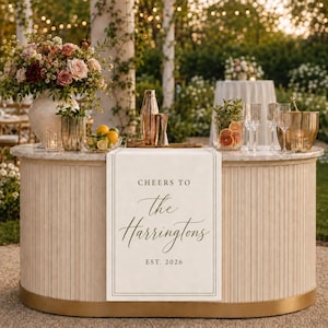 May include: A cream-colored table runner featuring the printed text 'CHEERS TO the Harringtons EST. 2026' in a dark serif and script font, draped over a curved, light-toned bar counter decorated with a floral arrangement, glassware, and a cocktail shaker.