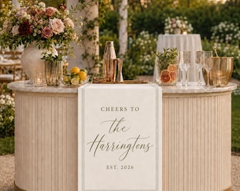 May include: A cream-colored table runner featuring the printed text 'CHEERS TO the Harringtons EST. 2026' in a dark serif and script font, draped over a curved, light-toned bar counter decorated with a floral arrangement, glassware, and a cocktail shaker.