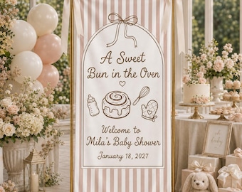 May include: A vertical banner with light brown and white vertical stripes, featuring a central arch design with a bow at the top. The text reads A Sweet Bun in the Oven, Welcome to Mila's Baby Shower, and January 18, 2027, with line art icons.