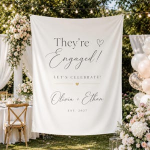 May include: A white banner with the text "They're Engaged!" in elegant script, along with "Let's Celebrate!" and the names "Olivia + Ethan" and the year "EST. 2027". The banner is decorated with a heart symbol and a gold heart.