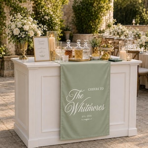 May include: A sage green table runner draped over a white bar, featuring white printed text that reads "CHEERS TO The Whitmores EST. 2026" in a decorative script font. The runner is positioned beneath three glass decanters and a framed sign.