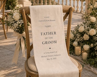 May include: A beige fabric memorial sign draped over a wooden chair, featuring the black printed text: a seat reserved from heaven, FATHER OF THE GROOM, in loving memory of Michael Thomas Anderson, May 18, 2025, with a small heart and leaf icon.