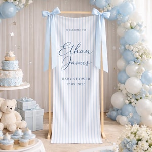 Op de afbeelding: Een verticale lichtblauw en wit gestreepte banner hangend aan een houten frame, met de tekst "WELCOME TO Ethan James BABY SHOWER 17.09.2026" in donkerblauw cursief en blokletters, versierd met lichtblauwe satijnen strikken aan de bovenhoeken.