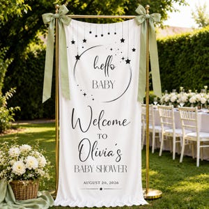 May include: A white vertical fabric banner suspended on a gold metal stand with sage green ribbon bows. The banner features black text reading "hello BABY", "Welcome TO Olivia's BABY SHOWER", and "AUGUST 20, 2026", decorated with a moon and stars.