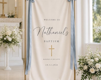 May include: A cream-colored fabric banner suspended on a gold metal stand, featuring the text "WELCOME TO Nathaniel's BAPTISM" with a gold cross icon and the date "10.12.2026". The banner is secured at the top with light blue satin ribbons.