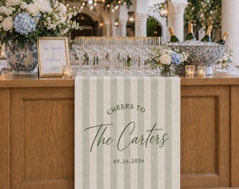 May include: A sage green and white striped table runner featuring the printed text Cheers To The Carters and the date 09.26.2026. The runner is displayed on a wooden bar with champagne flutes, a floral arrangement, and a framed sign reading Sip Sparkle & Celebrate.