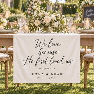 May include: A cream-colored fabric table runner featuring the black printed text We love because He first loved us, 1 John 4:19, EMMA & NOAH, and 06.14.2027. The runner is draped over a wooden table with a floral centerpiece and glassware.