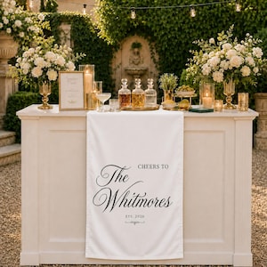 May include: A white fabric table runner featuring the black printed text 'CHEERS TO The Whitmores ETS. 2026' draped over a white bar counter. The counter is decorated with glass decanters, floral arrangements in gold vases, and lit candles.