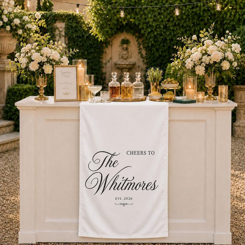May include: A white fabric table runner featuring the black printed text 'CHEERS TO The Whitmores ETS. 2026' draped over a white bar counter. The counter is decorated with glass decanters, floral arrangements in gold vases, and lit candles.