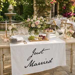 May include: A white table runner featuring the black text Just Married, draped over a wooden table set with a floral centerpiece, a platter of grapes and cheese, a cake stand with cupcakes, a wine bottle in an ice bucket, and white plates.
