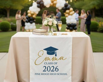 May include: A white graduation table runner with the name "Emma", a graduation cap, and the text "Class of 2026" and "Pine Ridge High School" in navy blue and gold glitter. The runner is on a beige tablecloth.