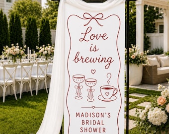 May include: A vertical white banner on a black metal stand featuring red line art of two stemmed glasses and a coffee cup. The text reads Love is brewing and MADISON'S BRIDAL SHOWER. A sheer white fabric drape hangs from the top of the frame.