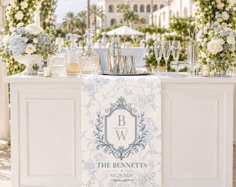 May include: A white table runner featuring a light blue floral pattern and a central crest with the initials BW, the text THE BENNETTS, and the date 05.20.2026. The runner is draped over a white bar counter set with glassware and a cocktail shaker.