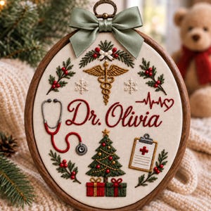 Puede incluir: Un bastidor de bordado circular de madera con el nombre Dr. Olivia en letra roja. El diseño incluye un caduceo dorado, un estetoscopio rojo, una línea de latido, un árbol de Navidad con regalos y ramas de acebo, rematado con un lazo de satén verde salvia.