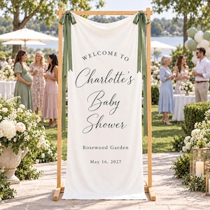 Op de afbeelding: Een verticaal wit stoffen spandoek hangend aan een licht houten frame, met de zwarte tekst "WELCOME TO Charlotte's Baby Shower Rosewood Garden May 16, 2027". Het spandoek is aan de bovenhoeken bevestigd met saliegroene linten in strikken.