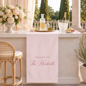 May include: A light pink and white vertical striped table runner featuring the text "CHEERS TO The Hartwells" in a dark pink script font. The runner is draped over a white counter with a glass decanter, a bowl of olives, and a champagne bucket.